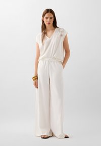 White short-sleeve V-neck top with floral embroidery, paired with loose white pants. Accessories include three wooden bangle bracelets.