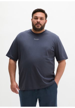 Mężczyzna z brodą, ubrany w granatową koszulkę Marc O'Polo i ciemne spodnie, stoi na jednolitym białym tle.