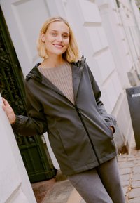 Sivi zip-up dukserica s kapuljačom, s elastičnim manžetama i glatkom teksturom, nosi se preko svjetlosive pletene džemperice, u kombinaciji s sivim hlačama.