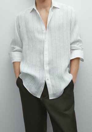 Personne portant une chemise blanche à manches longues boutonnée avec de fines rayures verticales et un pantalon foncé, les mains dans les poches, sur un fond uni.