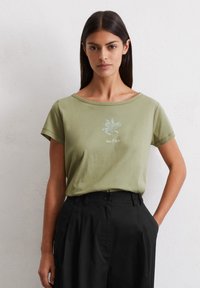 Olivenfarbendes T-Shirt mit kurzen Ärmeln und Rundhalsausschnitt, das ein florales Grafikdesign und Text in einer helleren Nuance zeigt. Getragen mit schwarzen Hosen.