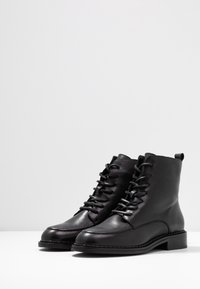 Botas de cuero negro hasta el tobillo con puntera redonda, cordones tradicionales y un tacón bajo en bloque. Presentan una textura suave y detalles de costura mínimos.