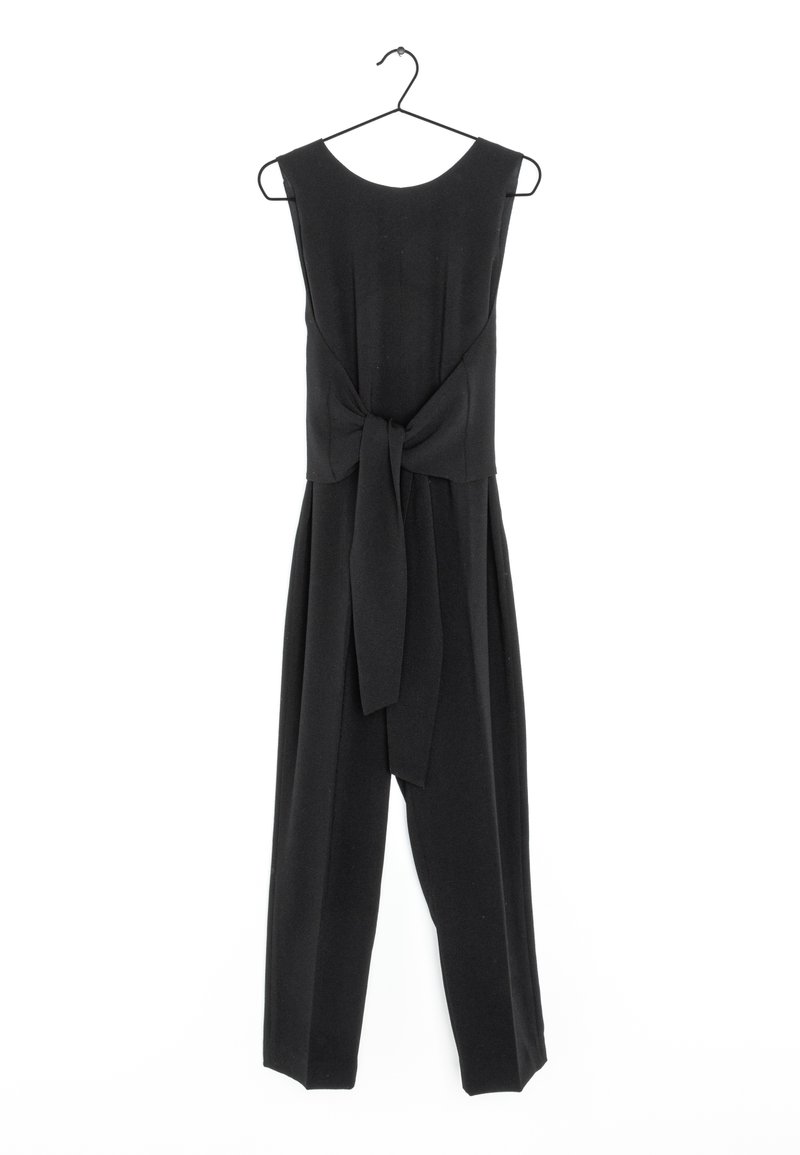 Combinaison noire sans manches avec jambes larges et ceinture en tissu nouée à la taille, suspendue à un cintre en métal noir sur fond blanc.