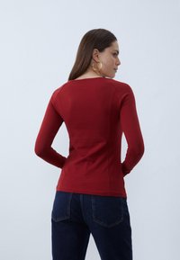 Red long-sleeve top, smooth texture, fitted design. Back view showing rounded neckline and seam detailing, paired with dark blue jeans.