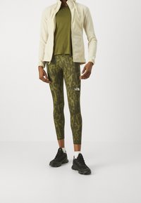 Leggings verde oliva con un patrón de camuflaje, combinados con una camisa verde oliva oscura y una chaqueta de forro polar cremosa con cremallera. Zapatillas deportivas negras.