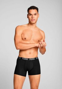 Black boxer briefs feature a thick, elastic waistband with "PUMA" logo. The fabric appears smooth with a fitted design. Model stands with hands clasped.