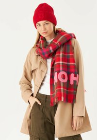Rote gestrickte Mütze, beiger Trenchcoat und karierter Schal in Rot-, Grün- und Rosatönen mit dem Text "HO". Fransen an den Schal-Enden.