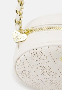 Kulatá kabelka krémové barvy s perforovaným logem, zlatým zipem a řetízkem, obsahující přívěsek ve tvaru srdce.