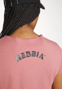 Camiseta rosa con cuello redondeado, que presenta el texto negro "NEBBIA" en un diseño semi-arco en la parte posterior. Tela texturizada visible.