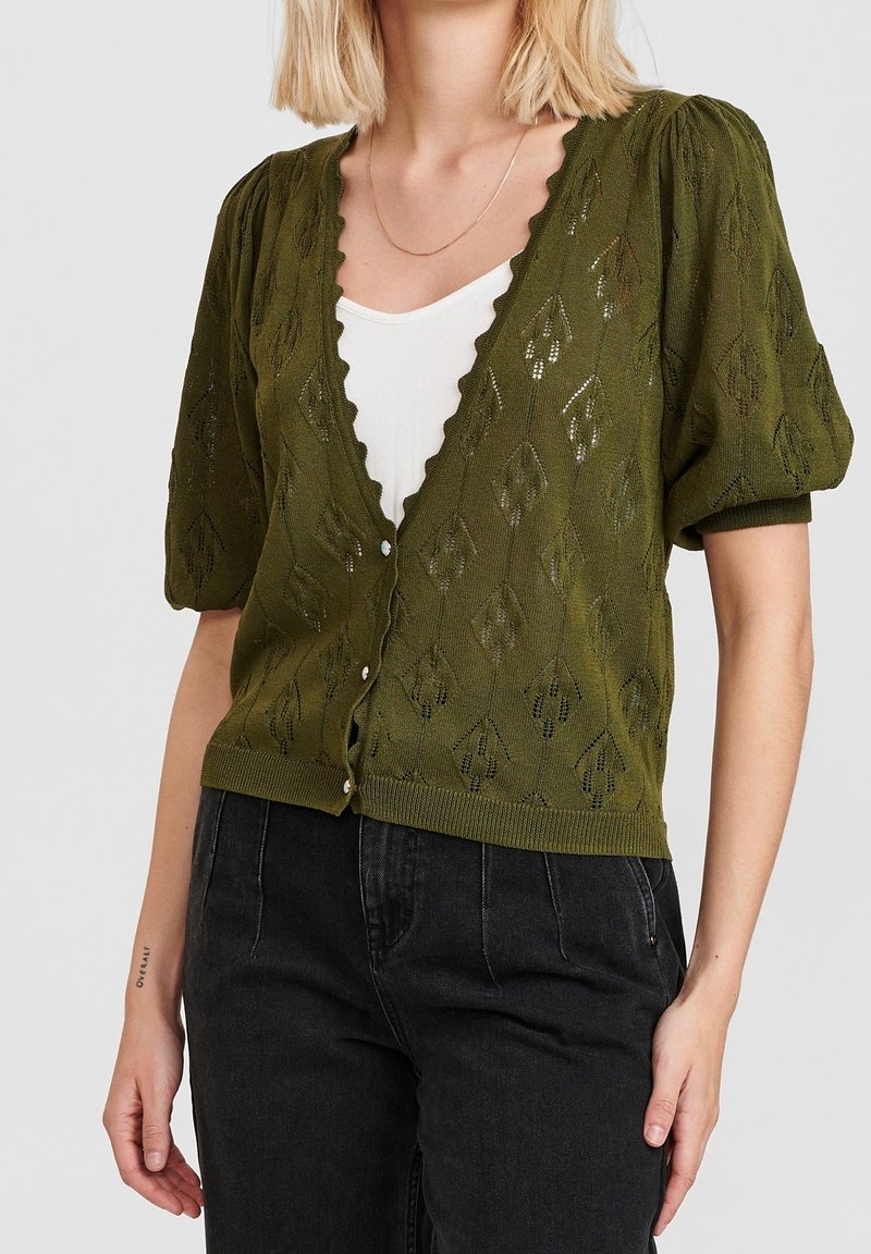 Femme portant un cardigan en tricot manches courtes vert olive avec des motifs de feuilles par-dessus un haut blanc et un jean noir.