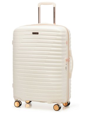 Kremowa walizka z twardą skorupą, wyposażona w cztery czarno-złote kółka, teleskopową rączkę, boczny uchwyt oraz poziomy teksturowany wzór.
