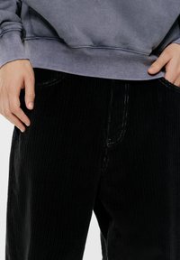Persoană îmbrăcată în pantaloni din catifea neagră și un hanorac gri, având mâinile odihnite aproape de buzunarele pantalonilor.