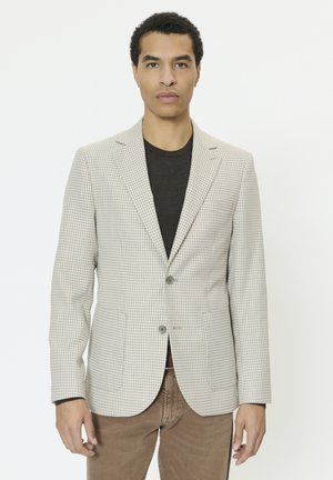 Homme portant un blazer beige à carreaux par-dessus une chemise noire et un pantalon marron, debout devant un fond blanc uni.