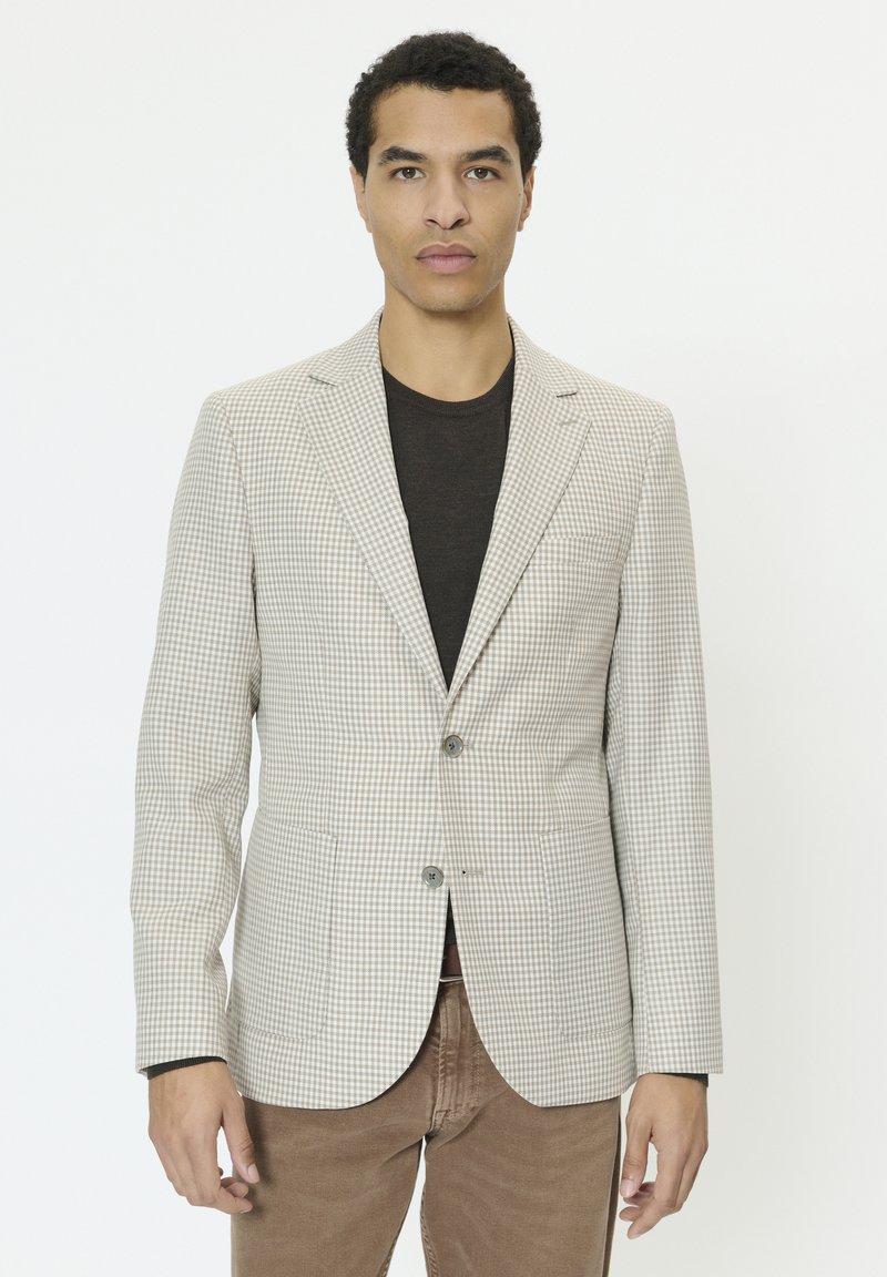 Mann trägt beige karierte Blazer über schwarzem Hemd und brauner Hose, steht vor schlichtem weißen Hintergrund.