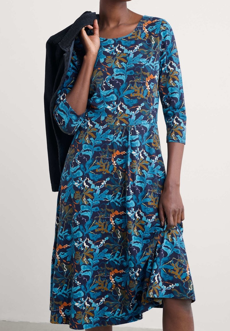 Femme portant une robe bleue à fleurs avec des manches trois-quarts, tenant une veste noire sur l'épaule et soulevant légèrement l'ourlet de la robe.