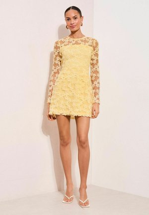 Žena v priliehavej žltej minišatách s priehľadnými dlhými rukávmi a kvetinovou čipkovou aplikáciou, doplnená bielymi sandálmi s odhalenými prstami a zlatými náušnicami kruhového tvaru.