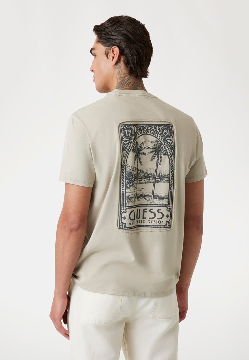 Lichtgrijze t-shirt met korte mouwen met een zwarte grafische print van palmbomen en een schilderachtig uitzicht met de tekst "GUESS AUTHENTIC DESIGN".
