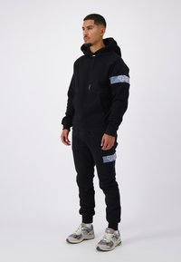 Zwarte hoodie met een grijze logo patch op de mouw, gecombineerd met bijpassende trainingsbroek met een grijze accentzak. Gemaakt met grijze sneakers.