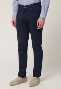 Pantalones de algodón azul marino de corte ajustado, con cinco bolsillos y cierre de botón. Llevados con zapatos tipo slip-on de color gris claro. Textura suave.