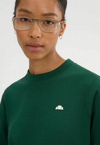 Sudadera verde de cuello redondo hecha de algodón, con un pequeño logo blanco en el pecho. El modelo lleva gafas y pendientes de aro.