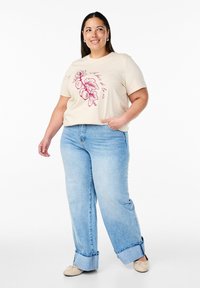 Beige t-shirt with a floral and text graphic in pink. Blue wide-leg jeans with a cuffed hem. Model wears flat shoes, standing with hands in pockets.