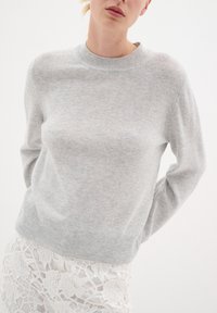 Pulover gri deschis realizat dintr-un material tricotat moale, având un decolteu și manșete din tricot cu nervuri, cu o croială dreaptă și o textură minimă.