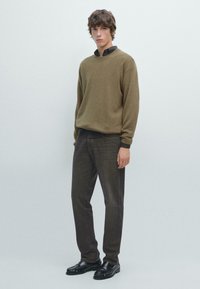 Pullover di lana marrone a collo tondo, abbinato a jeans dritti marrone scuro e mocassini neri, su uno sfondo grigio chiaro.
