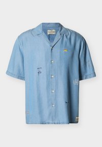 Ανοιχτό μπλε πουκάμισο με κοντά μανίκια και κουμπιά, με μικρές κεντημένες αφηρημένες φιγούρες προσώπων, ένα κίτρινο έμβλημα κορώνας και επιθέματα στο εμπρόσθιο μέρος και στα μανίκια.