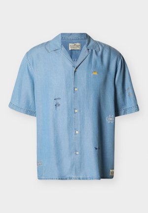Camicia celeste a maniche corte con bottoni, ricamata con piccoli volti astratti, un emblema di corona gialla e toppa sul davanti e sulle maniche.