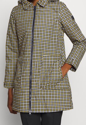 Femme portant une veste à carreaux jaunes, noirs et blancs avec fermeture éclair à l'avant et poche plaquée, main dans la poche, sur fond uni.