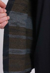 Mano sosteniendo una chaqueta abierta que muestra un forro de tela tejida a rayas marrón y gris por dentro.