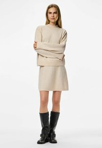 Beige stickad tröja med vida ärmar, rund halsringning och matchande kjol, kombinerad med knähöga svarta läderstövlar med spännen.