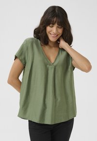 Blouse en soie verte avec un décolleté en V, manches courtes et coupe décontractée. Texture lisse avec un éclat subtil. Portée avec un pantalon noir.