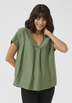 Blouse en soie verte avec un décolleté en V, manches courtes et coupe décontractée. Texture lisse avec un éclat subtil. Portée avec un pantalon noir.