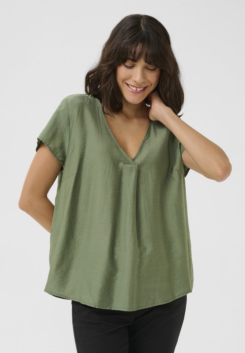 Blouse en soie verte avec un décolleté en V, manches courtes et coupe décontractée. Texture lisse avec un éclat subtil. Portée avec un pantalon noir.