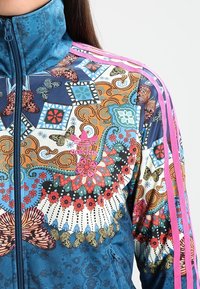 Blue zip-up jacket with multicolored floral and butterfly patterns; features pink stripes on shoulders and intricate designs throughout.
