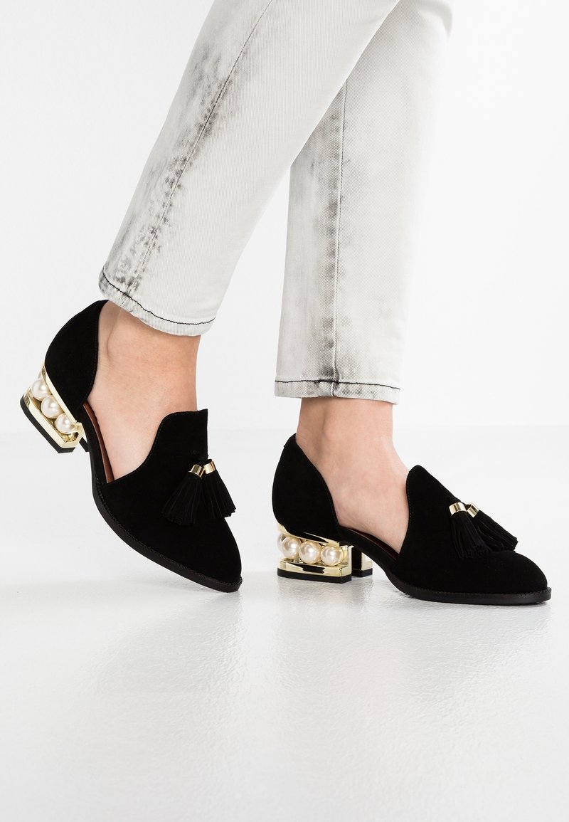 Chaussures en daim noir à talon bas, ornées d'un accent doré brillant et de perles factices, surmontées d'un détail à pampille noire à l'avant.