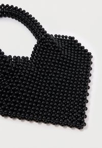 Μαύρη τσάντα χειρός με στρογγυλή λαβή και δομημένο, γωνιώδες κάτω μέρος. Η επιφάνεια έχει μια λεία υφή από ομοιόμορφες γυαλιστερές χάντρες.