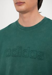 Zelené bavlnené tričko s textúrovým, vyšívaným logom "adidas" na prednej strane. Okrúhly výstrih a krátke rukávy, hladká textúra látky.