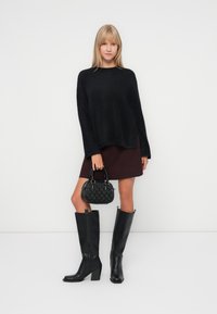 Pull en laine noir et duveteux, jupe bordeaux, bottes en cuir noir à hauteur de genou avec un talon block, et sac noir texturé avec de petites poignées.