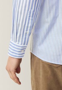 Gestricktes Hemd in Hellblau und Weiß, mit Knopfmanschetten, einem spitzen Kragen und glattem Stoff. Kombiniert mit braunen Cordhosen.