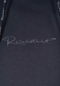 Tessuto nero con una superficie liscia, con un logo cucito in contrasto "Reichertstadt™" in argento nella sezione inferiore destra.