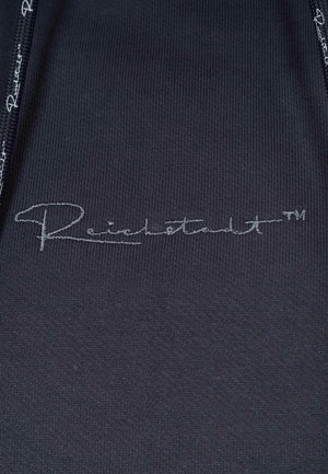Tissu noir avec une texture lisse, arborant un logo brodé contrastant "Reichertstadt™" en argent dans la section inférieure droite.