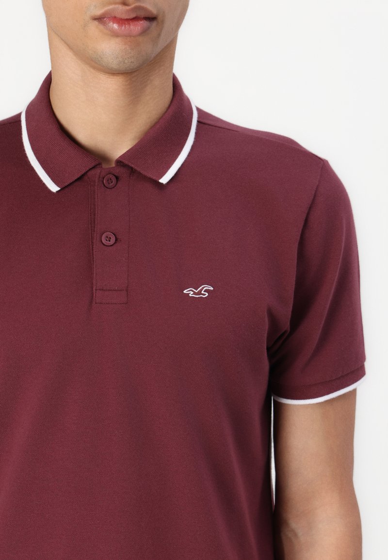 Polo bordeaux avec une texture unie, deux rayures blanches sur le col et un logo brodé sur la poitrine gauche.
