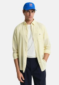 Geel gestreepte overhemd met lange mouwen, gecombineerd met een witte t-shirt en marineblauwe broek. Blauwe pet met logo aan de voorkant.
