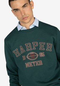 Felpa verde con grafica "HARPER RUGBY TEAM 1985" in lettere chiare e scure. Design a girocollo con texture a coste e dettagli a contrasto.