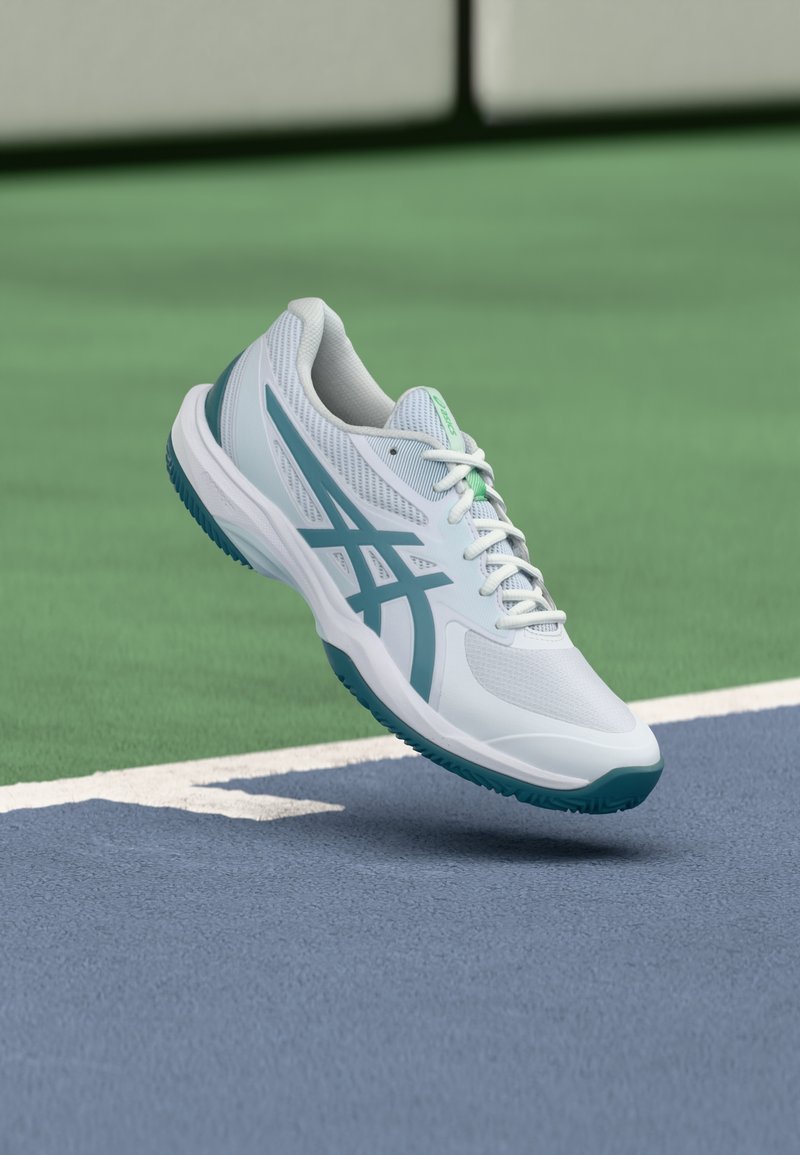 Witte sportschoen met turquoise accenten, een bovenwerk van mesh en een patroonprofiel op de zool, zwevend boven een blauwe sportvloer. Voorzien van veterluiting en gevoerde kraag.