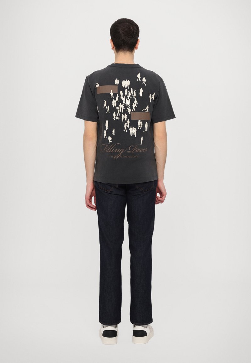 Dark gray short-sleeve t-shirt featuring a graphic of white outlined figures and "Filling Pieces" text on the back. Paired with dark denim jeans.