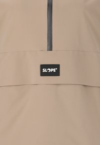 Beige vedenkestävä takki, jossa on edessä vetoketju ja musta "SLOPE" -logopatchi. Sileä pinta ja minimalistinen muotoilu.
