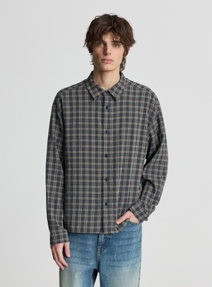 Młody mężczyzna z falującymi brązowymi włosami, ubrany w niebiesko-beżową kraciastą koszulę zapinaną na guziki oraz jasnoniebieskie dżinsy, stojący na jednolitym tle.